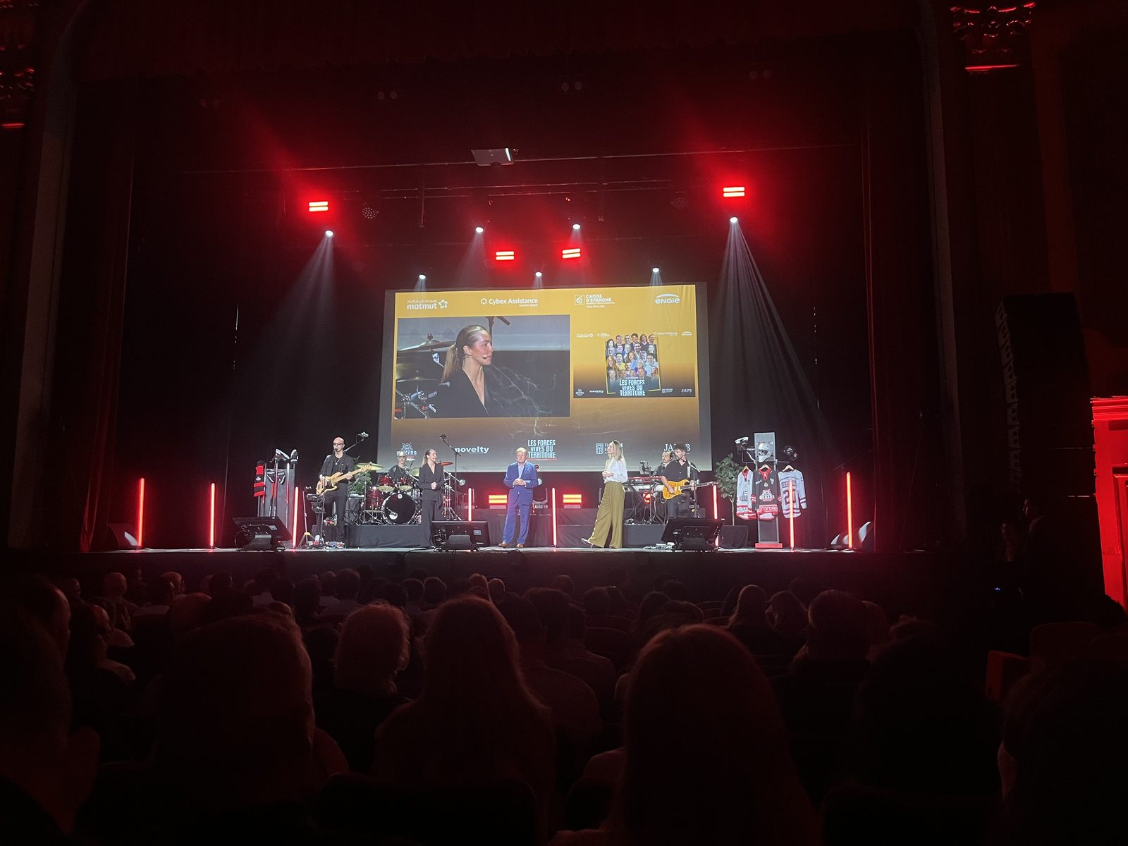 Scène du Théâtre Fémina pendant les Forces vives du Territoire : un groupe de musique, deux animateurs et une intervenante en visioconférence sur le grand écran